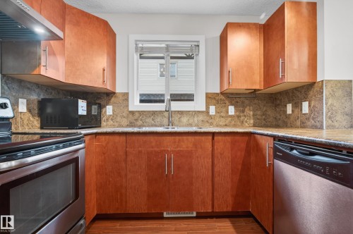 Kitchen featuring wood-finish cabinetry with brushed nickel hardware, a stainless steel range with overhead hood, a stainless steel dishwasher, a window above the sink, and granite-look countertops with a matching backsplash - 48 8716 179 Avenue, Edmonton, AB 