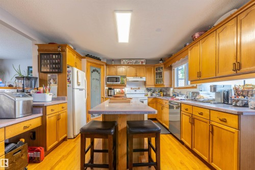Kitchen featuring light wood cabinetry, a central island with countertop seating, wood-finish flooring, a white refrigerator, and a built-in dishwasher - 8902 107 Avenue, Morinville, AB 