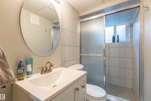 Bathroom featuring a single vanity with a white countertop and basin, a round wall mirror, a toilet, a shower with a sliding glass door, and white tiled walls - 33 Andrew Crescent, St. Albert, AB 