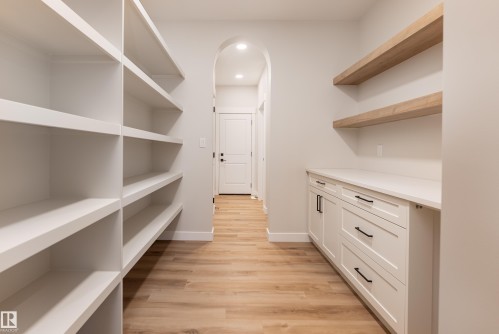 Dedicated storage area featuring extensive built-in shelving, white cabinetry with black hardware, a light-toned countertop, wood-finish flooring, and arched doorway - 115 Garneau Gate, Spruce Grove, AB 