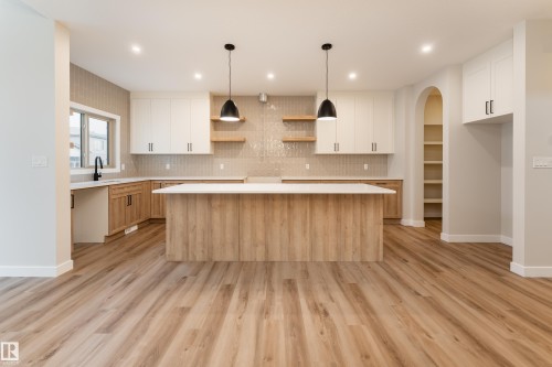 Open-concept kitchen featuring wood-finish flooring, a central island with a white countertop, two-tone cabinetry, a textured tile backsplash, and an arched pantry alcove with shelving - 115 Garneau Gate, Spruce Grove, AB 