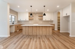Open-concept kitchen featuring wood-finish flooring, a central island with a white countertop, two-tone cabinetry, a textured tile backsplash, and an arched pantry alcove with shelving - 
