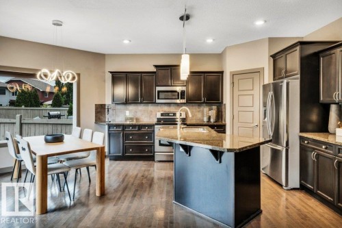 Open-concept kitchen and dining area featuring wood-finish flooring, dark wood cabinetry, a granite-top island, stainless steel appliances, and a tiled backsplash - 1107 Chahley Crest, Edmonton, AB 