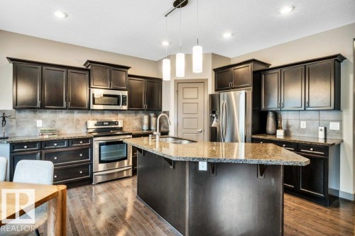 Kitchen featuring dark wood-finish cabinetry, stainless steel appliances, granite countertops, a tiled backsplash, and wood-finish flooring - 1107 Chahley Crest, Edmonton, AB 