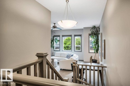 Staircase with painted wood banister leading to an upper landing featuring carpeted flooring, a contemporary pendant light fixture, and multiple windows with grey trim - 1107 Chahley Crest, Edmonton, AB 