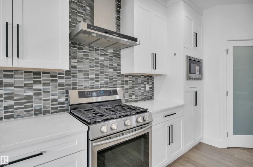 Gourmet kitchen featuring a stainless steel range with a coordinating hood, grey mosaic tile backsplash, white shaker cabinetry with matte black hardware, white countertops, and a built-in microwave - 3898 Robins Crescent, Edmonton, AB 