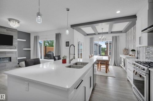 Open-concept kitchen and living area featuring a large kitchen island with a double basin sink, stainless steel appliances, coffered ceilings in the dining area, a tiled fireplace, and wood-finish flooring - 3898 Robins Crescent, Edmonton, AB 