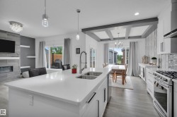Open-concept kitchen and living area featuring a large kitchen island with a double basin sink, stainless steel appliances, coffered ceilings in the dining area, a tiled fireplace, and wood-finish flooring - 