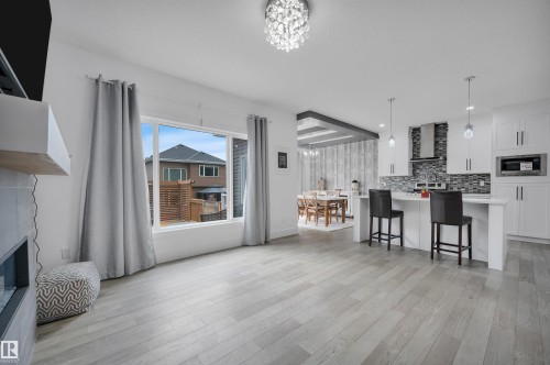 Open-concept interior featuring light wood-finish flooring, a modern kitchen with a white island and pendant lighting, white cabinetry, a mosaic tile backsplash, and a contemporary fireplace - 3898 Robins Crescent, Edmonton, AB 