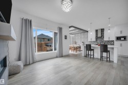 Open-concept interior featuring light wood-finish flooring, a modern kitchen with a white island and pendant lighting, white cabinetry, a mosaic tile backsplash, and a contemporary fireplace - 