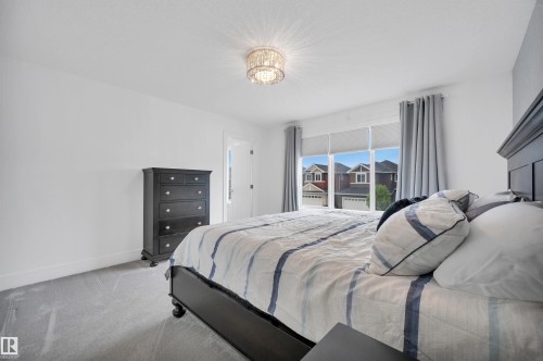 Bedroom featuring light gray carpeting, a crystal-style ceiling light fixture, and a large window with roller shades and gray drapes - 3898 Robins Crescent, Edmonton, AB 