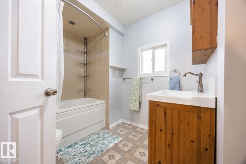 Bathroom featuring a wood-finish vanity with an integrated white sink, a window, a tiled shower/tub combination, and patterned floor tiles - 17932 93 Avenue, Edmonton, AB 