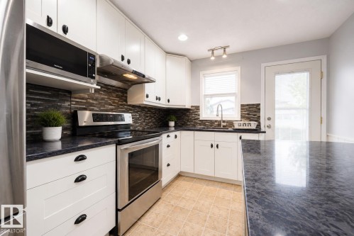 Modern kitchen featuring white cabinetry with black hardware, stainless steel appliances, dark countertops, and a mosaic tile backsplash - 17932 93 Avenue, Edmonton, AB 