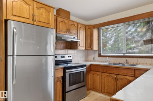 Kitchen featuring wood cabinetry, stainless steel appliances, a double basin sink, and a tiled backsplash - 13920 64 Street, Edmonton, AB 