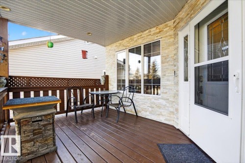 Covered porch featuring wood-finish decking, stone-faced exterior wall accents, a white storm door with decorative glass, white vinyl siding, and a lattice privacy screen - 5019 48 Avenue, Chipman, AB - Outdoor With Deck Patio Veranda With Exterior
