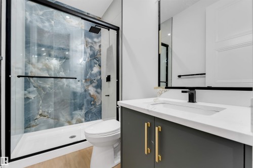 Spacious bathroom featuring a glass-enclosed shower with blue-toned marble-look wall tiling, a white vanity with gold-toned hardware, a rectangular undermount sink, and wood-finish flooring - 131 24 Street, Edmonton, AB 