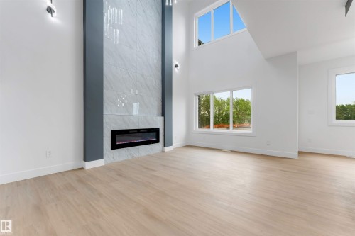 Double-height ceiling living space featuring a floor-to-ceiling accent wall with an electric fireplace, light wood-finish flooring, and multiple large windows - 131 24 Street, Edmonton, AB 