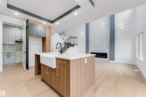 Open-concept living space featuring a central kitchen island with a farmhouse sink, wood-finish flooring, a modern fireplace with a tiled surround, and a glass-paneled staircase - 131 24 Street, Edmonton, AB 