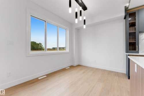 Bright interior space featuring a large three-panel window, light wood-finish flooring, a contemporary linear chandelier, white baseboards, and a partial view of built-in cabinetry with glass inserts - 131 24 Street, Edmonton, AB 