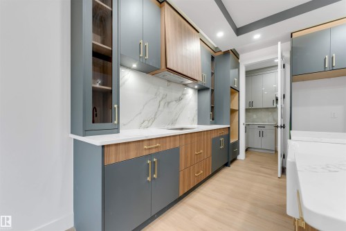 Modern kitchen featuring two-tone cabinetry with gold-tone hardware, white stone countertops, a large format marbled backsplash, and light wood-finish flooring - 131 24 Street, Edmonton, AB 