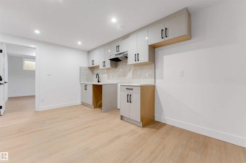 Contemporary kitchen area featuring light wood-finish flooring, recessed lighting, white cabinetry with matte black hardware, and a tile backsplash - 131 24 Street, Edmonton, AB 