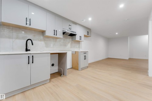 Open-concept living space featuring light wood-finish flooring, recessed lighting, and a contemporary kitchenette with light gray cabinetry, a white countertop, and a black gooseneck faucet - 131 24 Street, Edmonton, AB 