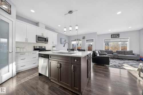 Open-concept living space featuring wood-finish flooring, a kitchen island with pendant lighting, white cabinetry, stainless steel appliances, and large windows - 2084 Graydon Hill, Edmonton, AB 