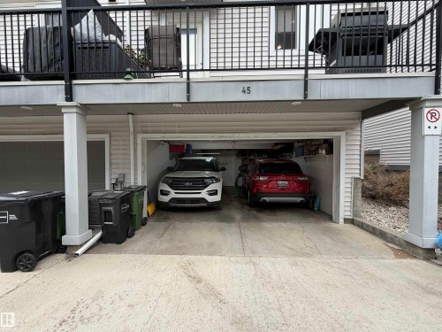 Two-car garage with overhead lighting, featuring a concrete driveway and an upper-level balcony with black railings - 45 6075 Schonsee Way, Edmonton, AB 