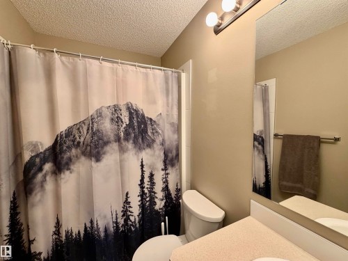 Bathroom featuring a shower with tiled surround, a toilet, a vanity with a light-colored countertop, a wall-mounted mirror, and overhead lighting - 45 6075 Schonsee Way, Edmonton, AB 