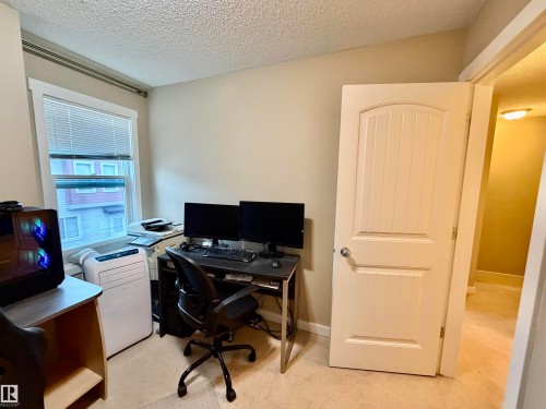 Carpeted room with a single window featuring white blinds and trim, a textured ceiling, and neutral-toned walls - 45 6075 Schonsee Way, Edmonton, AB 