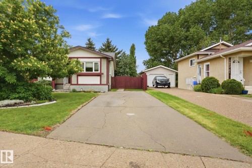 Property exterior featuring a light-colored facade with contrasting trim, a gabled roofline, mature trees, and a spacious driveway leading to a detached garage - 1008 61 Street, Edmonton, AB 