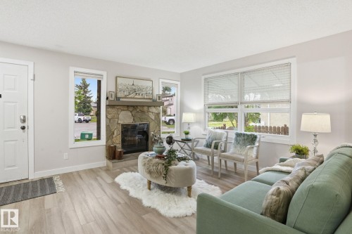 Living area featuring a stone-clad fireplace, light wood-finish flooring, multiple windows with blinds, a white entry door, and light-toned wall paint - 1008 61 Street, Edmonton, AB 