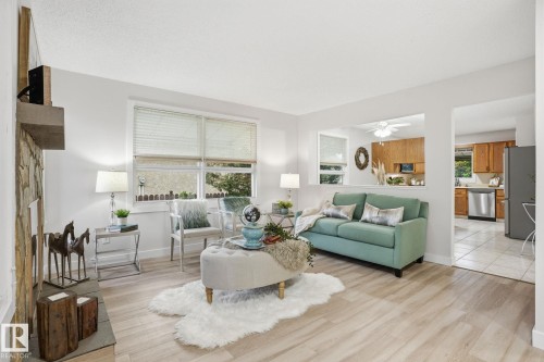 Bright living area featuring wood-finish flooring, a stone fireplace, large windows, and a pass-through opening to a kitchen with wood cabinetry and stainless steel appliances - 1008 61 Street, Edmonton, AB 