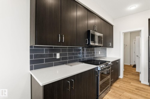 Modern kitchen featuring dark wood-finish cabinetry, light-toned countertops, a full subway tile backsplash, stainless steel appliances, and wood-look flooring - 20011 26, Edmonton, AB 