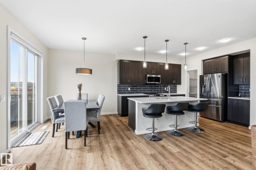 Open-concept kitchen and dining area featuring wood-finish flooring, dark wood cabinetry, stainless steel appliances, a kitchen island with seating, and sliding glass doors - 20011 26, Edmonton, AB 
