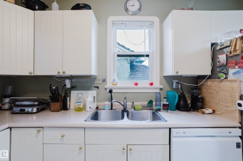 Kitchen area featuring white cabinetry with gold-toned hardware, a double basin stainless steel sink, and a white dishwasher - 11158 65 Street, Edmonton, AB 