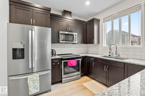 Modern kitchen featuring dark wood cabinetry, stainless steel appliances, speckled stone countertops, and a subway tile backsplash - 20227 17Ave, Edmonton, AB 