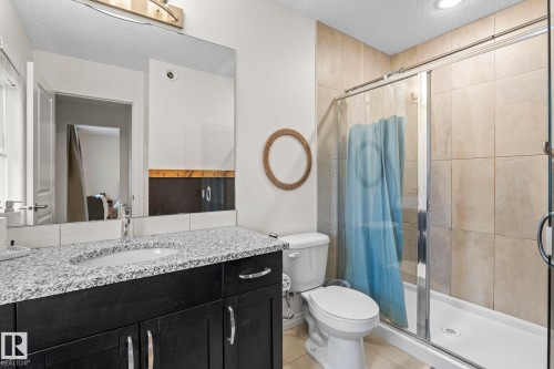 Bathroom featuring a dark wood-finish vanity with a speckled countertop, an undermount sink, a large wall mirror, and a glass-enclosed shower with tiled walls - 20227 17Ave, Edmonton, AB 
