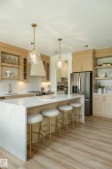 Spacious kitchen featuring a large waterfall island with integrated seating, light wood-finish cabinetry, stainless steel appliances, open shelving, and recessed lighting - 