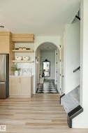 Interior hallway featuring wood-finish flooring, a built-in light wood cabinetry with open shelving and white countertops, a stainless steel refrigerator, and an arched doorway leading to a black and white checkered floor - 