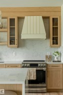 Kitchen featuring a paneled range hood, stainless steel range, light wood-finish cabinetry, glass-front upper cabinets, and light-toned countertops with matching backsplash - 