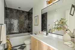 Bathroom featuring a white tub with dark subway tile surround, a light wood-finish vanity with a white countertop, a rectangular undermount sink, and matte black fixtures - 