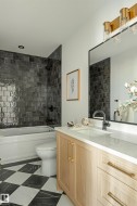 Bathroom featuring a light wood-finish vanity with a white countertop and rectangular sink, black and white checkered floor tiles, a built-in tub with dark subway tile surround, and a rectangular mirror with an overhead brass light fixture - 