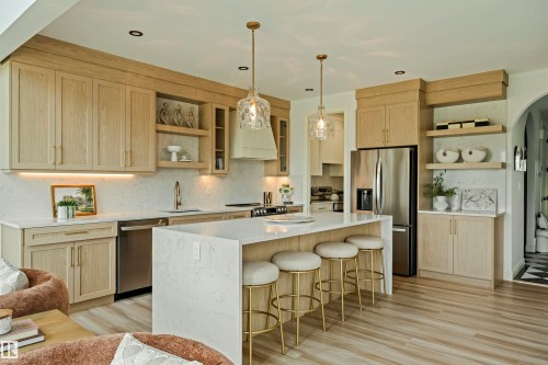 Open-concept kitchen featuring light wood-finish cabinetry, a large central island with seating, stainless steel appliances, and sleek pendant lighting - 3329 Chickadee Drive, Edmonton, AB 