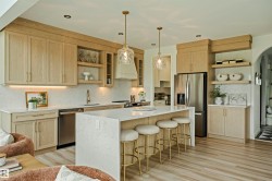 Open-concept kitchen featuring light wood-finish cabinetry, a large central island with seating, stainless steel appliances, and sleek pendant lighting - 