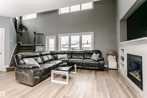Spacious living area featuring light wood-finish flooring, a white mantel fireplace with built-in shelving, large windows with white trim, a high ceiling, and a dark wood staircase with black railings - 21807 97 Avenue, Edmonton, AB 