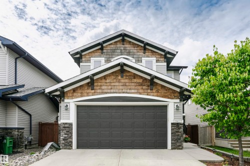 Craftsman-style facade featuring gray siding, natural wood shingles, dark gray garage door, stone pillar accents, and decorative roofline brackets - 21807 97 Avenue, Edmonton, AB 