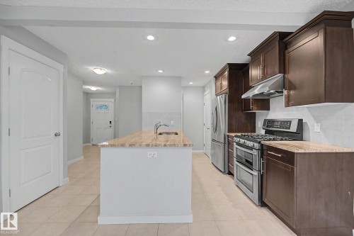 Kitchen featuring a central island with an undermount sink, dark wood cabinetry, stainless steel appliances, neutral tile flooring, and recessed lighting - 16810 53 Street, Edmonton, AB 