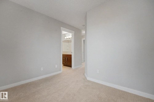 Neutral-toned wall paint and carpeting throughout, with a clear doorway leading to a bathroom featuring a wooden vanity, light countertop, and wall-mounted light fixture - 16810 53 Street, Edmonton, AB 