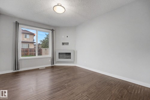 Spacious room featuring wood-finish flooring, a contemporary wall-mounted fireplace, and a large window with privacy curtains - 16810 53 Street, Edmonton, AB 